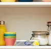 A shelf with various items including a yellow water bottle, colorful bowls, and jars of ingredients, with a pack of gum in the foreground. Produktbild-Vorschau