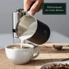 A hand is pouring frothed milk from a black milk frothing pitcher into a white coffee cup, with a coffee grinder in the background. Produktbild-Vorschau