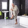 An elderly man in a bathrobe is applying lotion to his leg while sitting on a shower stool in a modern bathroom. Produktbild-Vorschau