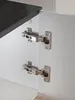 Close-up view of cabinet hinges on a white cabinet door, with a dark wood shelf visible at the top. Produktbild-Vorschau