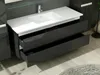 A modern bathroom vanity with a white countertop and sink, featuring two open black drawers with a textured circular pattern. The background includes a gray tiled wall and a wooden floor. Produktbild-Vorschau