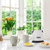 A white KitchenAid kettle sits on a countertop next to two mugs and a plate of cookies, with a vase of tulips and a potted plant in the background, all viewed through a window with greenery outside. Produktbild-Vorschau