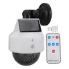 A solar-powered surveillance camera dummy with a remote control, screws, and wall plugs, displayed on a white background. Produktbild-Vorschau