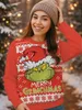 A woman wearing a Grinch-themed Christmas sweater and a beanie, smiling and posing outdoors in a snowy setting with blurred Christmas lights in the background. Náhľad obrázku produktu