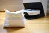 A white paper bag with a green logo and text sits on a wooden floor in front of a white robotic vacuum cleaner docked in its base. Produktbild-Vorschau