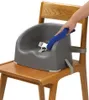 A person's hand is wiping a gray booster seat attached to a wooden chair with a blue cloth. Produktbild-Vorschau