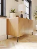 A wooden cabinet with a chevron pattern on its doors, supported by black metal legs, is placed on a herringbone wooden floor. A white lamp with a black base and a potted plant are on top of the cabinet. Produktbild-Vorschau