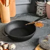 A black cast iron frying pan with a tag tied to its handle sits on a speckled gray countertop. In the background, a bamboo utensil holder with wooden spoons, a decorative jar, and a pink mug are visible. Produktbild-Vorschau