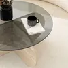 A close-up view of a round coffee table with a glass top, a book, and a mug on it, placed on a fluffy white rug. Produktbild-Vorschau