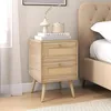 A wooden nightstand with rattan drawer fronts is placed next to a bed. Produktbild-Vorschau
