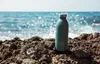 A teal water bottle sits on rocks by the ocean. Produktbild-Vorschau