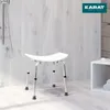 A white shower stool with adjustable legs is placed on a light wooden floor in a modern bathroom setting, with a shower head visible on the left and a white storage cube with towels on the right. Podgląd zdjęcia produktu