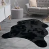 A black faux fur rug is placed on a wooden floor in a living room setting, next to a gray sofa and a round ottoman. Produktbild-Vorschau