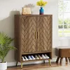 A wooden shoe cabinet with a herringbone pattern on the doors, gold handles, and gold legs. It has an open shelf at the bottom displaying several pairs of shoes. A handbag and a vase with yellow flowers are on top of the cabinet. A potted plant is to the l Náhľad obrázku produktu