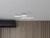 A modern ceiling light fixture with two curved LED light bars is mounted on a white ceiling above a dark air conditioning unit and a wooden paneled wall. Náhled obrázku produktu