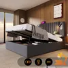 A modern bedroom with a grey upholstered bed with its storage compartment open, showing items inside. Large windows with a city view are in the background. Náhľad obrázku produktu