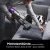A person is using a cordless vacuum cleaner with a purple accent on a couch where a dog is lying. Produktbild-Vorschau