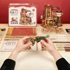 A flat lay of a ROKR 3D-Holz-Puzzle Dream Gift Factory kit, showing the box, unassembled wooden pieces, and a partially assembled model. Hands are holding a small, assembled component of the puzzle. Podgląd zdjęcia produktu