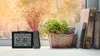 A projection alarm clock with weather forecast is displayed on a windowsill next to books and a plant, with rain visible on the window. Produktbild-Vorschau