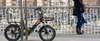 A person stands on a bridge next to an e-bike, with a city skyline in the background. Produktbild-Vorschau
