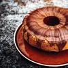 A bundt cake with chocolate drizzle on a brown plate, set against a dark, speckled background. Anteprima dell’immagine prodotto