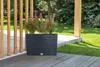 A dark gray rectangular planter with green and red-leafed plants sits on a wooden deck next to a wooden fence. Produktbild-Vorschau