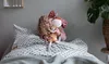 A young girl is lying in bed, holding a doll and a smaller stuffed animal. Produktbild-Vorschau