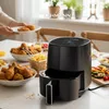 A table laden with fried chicken and fries, with a person placing more chicken onto a plate, and a Lehmann Home air fryer prominently displayed. Produktbild-Vorschau
