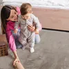 A mother is helping her baby stand up on a soft carpet next to a bed. Produktbild-Vorschau