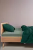 A bed with green bedding and a pillow against a light pink wall. Produktbild-Vorschau