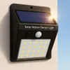 A black solar-powered motion sensor light is mounted on a light-colored wall, with a bright sun shining in the upper right corner and a blue sky in the background. Produktbild-Vorschau