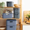 Storage boxes are neatly stacked on wooden shelves, with some greenery and kitchenware in the background. Produktbild-Vorschau
