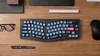 A top-down view of a split ergonomic keyboard on a wooden desk, with a mouse, glasses, and a pen. Anteprima dell’immagine prodotto