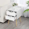 A white nightstand with two drawers is placed next to a white sofa, with a bouquet of flowers and a teacup on top. Produktbild-Vorschau