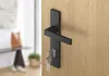 A black door handle with keys inserted into the lock, mounted on a wooden door. The background shows a blurred interior of a hallway with a table and a bag. Produktbild-Vorschau