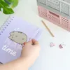 A hand is holding a purple notebook with a Pusheen cat design on it, with a pencil and binder clips on a white desk. Náhľad obrázku produktu