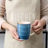 A person wearing an apron holds a blue ceramic mug. Náhled obrázku produktu