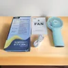 A handheld fan, its user manual, packaging, and charging cable are laid out on a wooden table. Produktbild-Vorschau