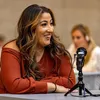 A woman is smiling while sitting at a table with a microphone in front of her. Náhled obrázku produktu