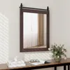 A mirror with a dark brown frame and a sliding barn door mechanism is mounted on a white paneled wall above a wooden console table. On the table, there are books, a candle, glasses, and a vase of pink flowers. Podgląd zdjęcia produktu