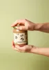 Hands holding a jar of hazelnut spread against a light green background. Produktbild-Vorschau
