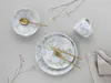 A flat lay of a dinnerware set with a floral pattern, including plates, a bowl, a mug, cutlery, and a napkin, arranged on a light gray background with decorative branches. Náhľad obrázku produktu