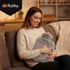 A woman is sitting on a couch, holding a gray plush hot water bottle. Náhled obrázku produktu