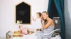 A mother and child are on a bed in a child's room. The mother is touching her ear, and the child is standing and looking away. Produktbild-Vorschau