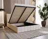 A beige upholstered bed frame with the mattress support mechanism open, revealing storage space underneath. The bed is placed on a rug in a room with wooden wall paneling and a bookshelf in the background. Náhled obrázku produktu