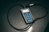 A digital thickness gauge with a cable and probe is laid out on a dark surface. Podgląd zdjęcia produktu
