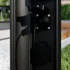 A close-up view of a black outdoor lamp post with two electrical outlets, one with a cord plugged in. Produktbild-Vorschau