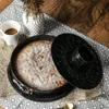A cake in a baking pan with a lid, next to a cup of coffee and a vase with dried flowers, on a patterned tablecloth. Anteprima dell’immagine prodotto