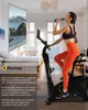 A woman is riding a stationary bike in a room, drinking from a bottle, with a large screen showing a mountain road in front of her. Produktbild-Vorschau