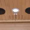 A close-up view of the ceiling of a wooden sauna, showing a light fixture, a temperature sensor, and two speakers. Produktbild-Vorschau